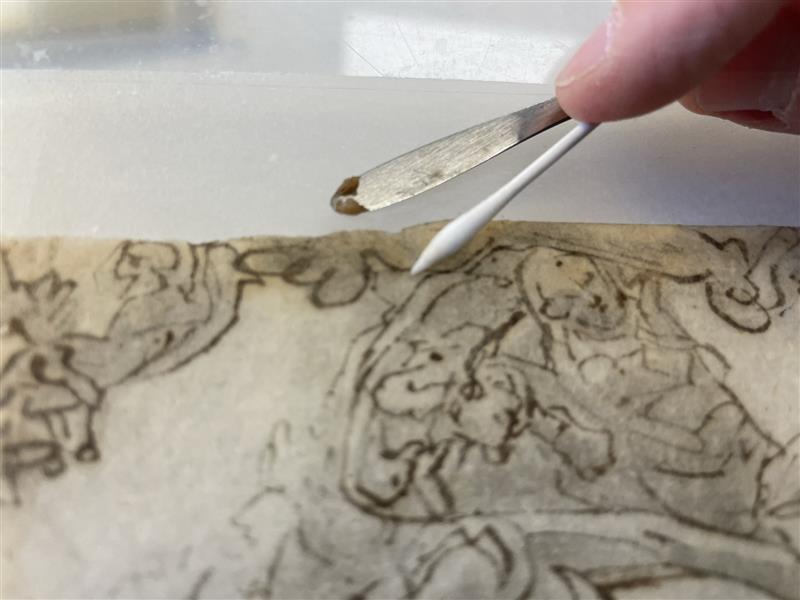 Close up of a work on paper utilizing iron gall ink, with a micro-spatula and a cotton swab hovering over the work.