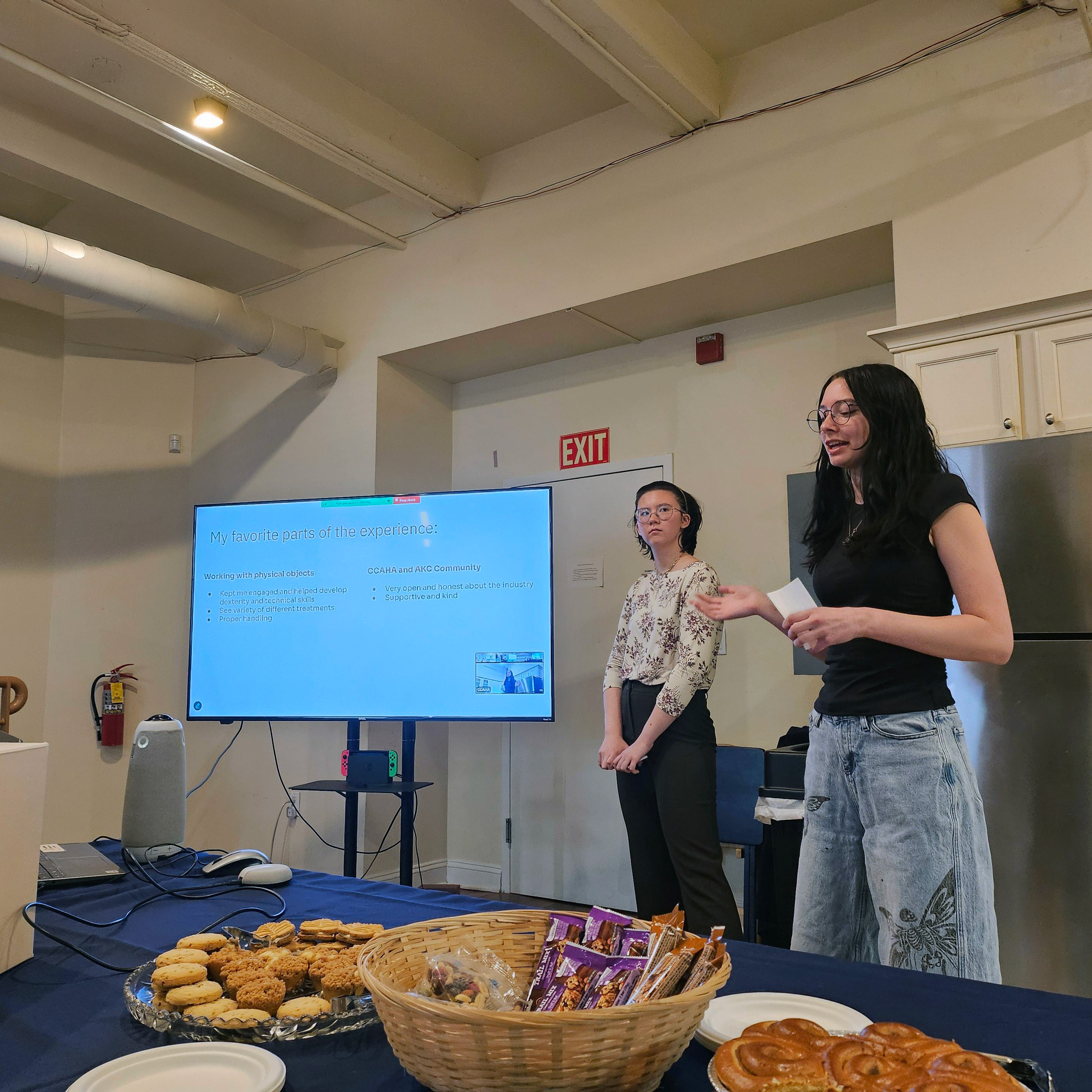 Two co-op students speaking in the center of the room with a tv monitor and table of refreshments.